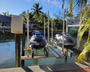Two jet skis are parked on a boat lift next to palm trees.
